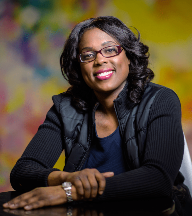 Studio portrait of Sandy Linda, hands crossed, smiling while looking at the camera.